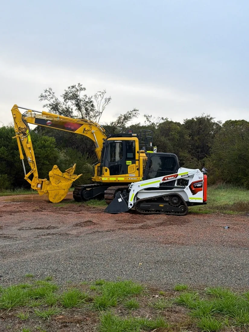 Komatsu excavator fitted with a tined rake bucket, standing on a cleared site alongside a tracked loader