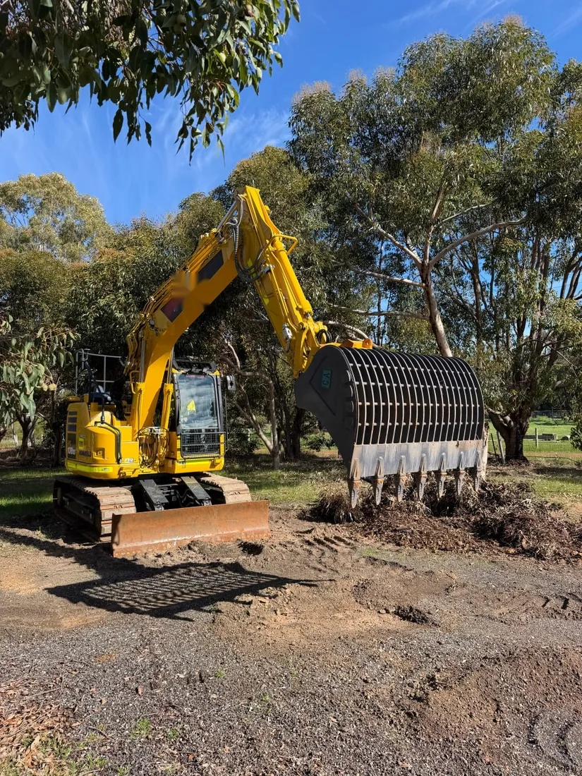Komatsu excavator fitted with a large steel screening bucket, parked beside a heap of screened material