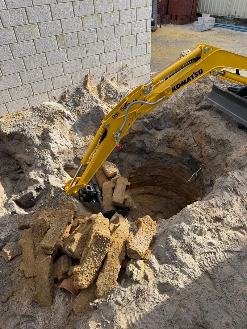 Mini excavator lifting limestone blocks clear of an excavation, safe working load stencilled on the boom