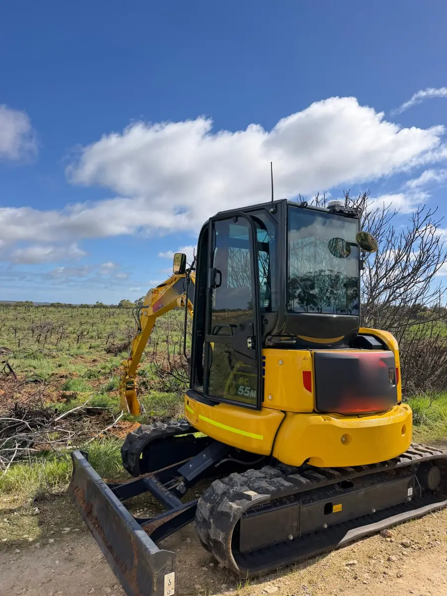 Komatsu mini excavator with a dozer blade standing at the edge of an old vineyard, bare vine rows still in the ground behind it