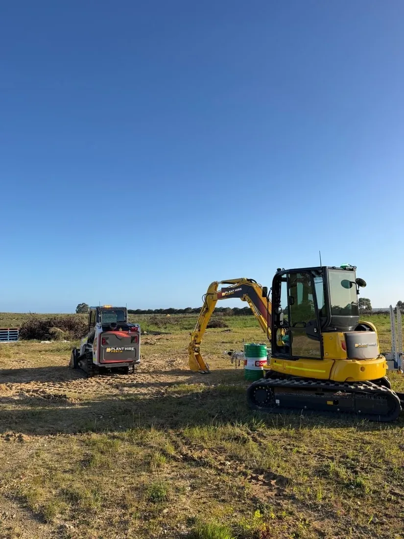 Bobcat track loader and Komatsu excavator standing on a cleared paddock, with pulled vineyard material heaped at the fence line